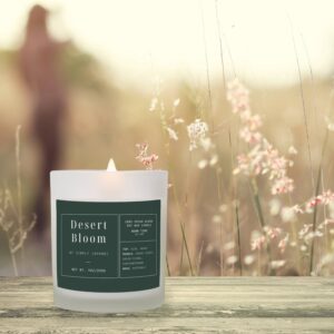 A lit scented candle labeled "Desert Bloom by Simply Japandi" sits on a wooden surface with blurred grass and a hazy figure in the background.