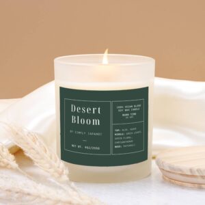 A lit white luxurious candle in a frosted glass jar labeled "Desert Bloom by Simply Japandi" on a beige background. The label lists its vegan soy wax, burn time of 50 hours, and notes of aloe, agave, green leaves, chrysanthemum, and patchouli.